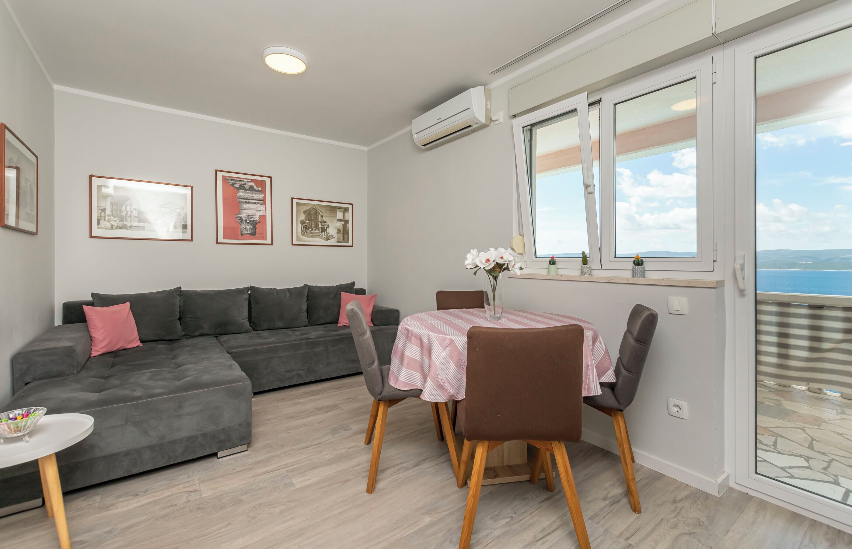 Un soggiorno moderno con un divano modulare grigio, un tavolo da pranzo con sedie e un balcone con vista sul mare.