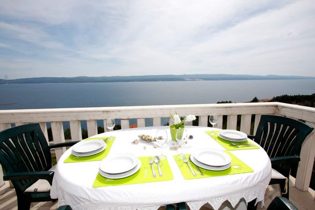 A round table set for four with white plates, green placemats, and silverware, overlooking a body of water.