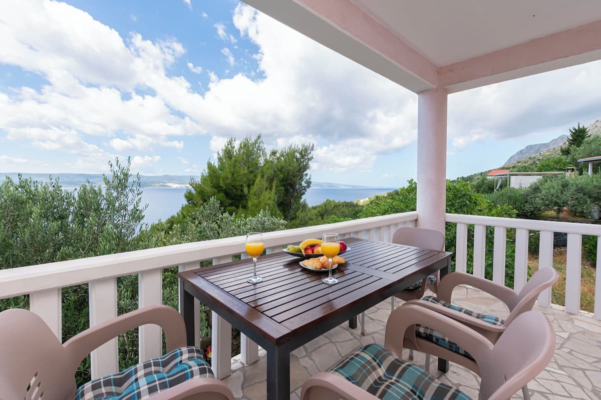 A wooden table with chairs on a balcony overlooking a scenic view of the sea and mountains.