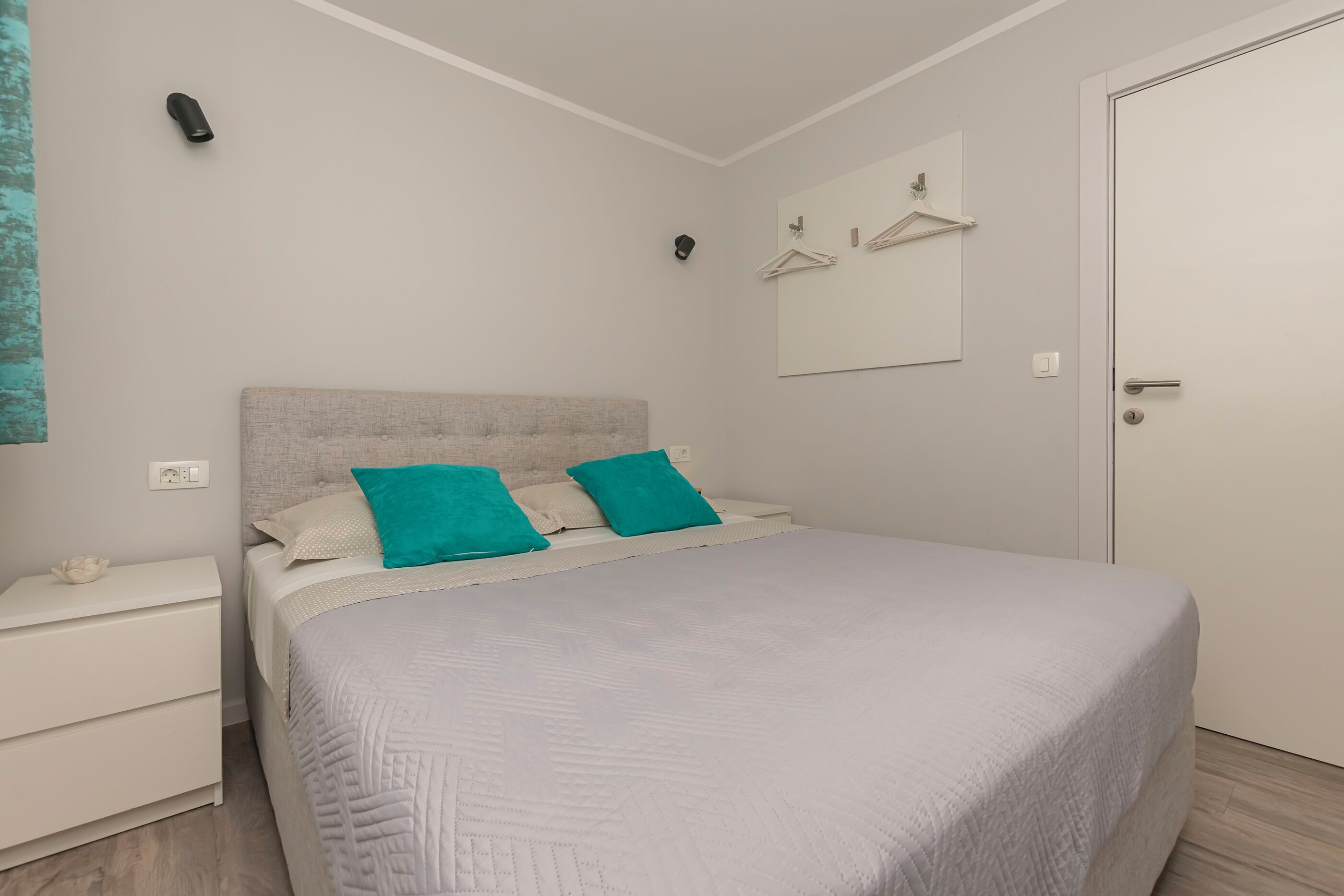 A bedroom with a bed, two pillows, a bedside table, a nightstand, a wall-mounted light, a mirror, and a door.