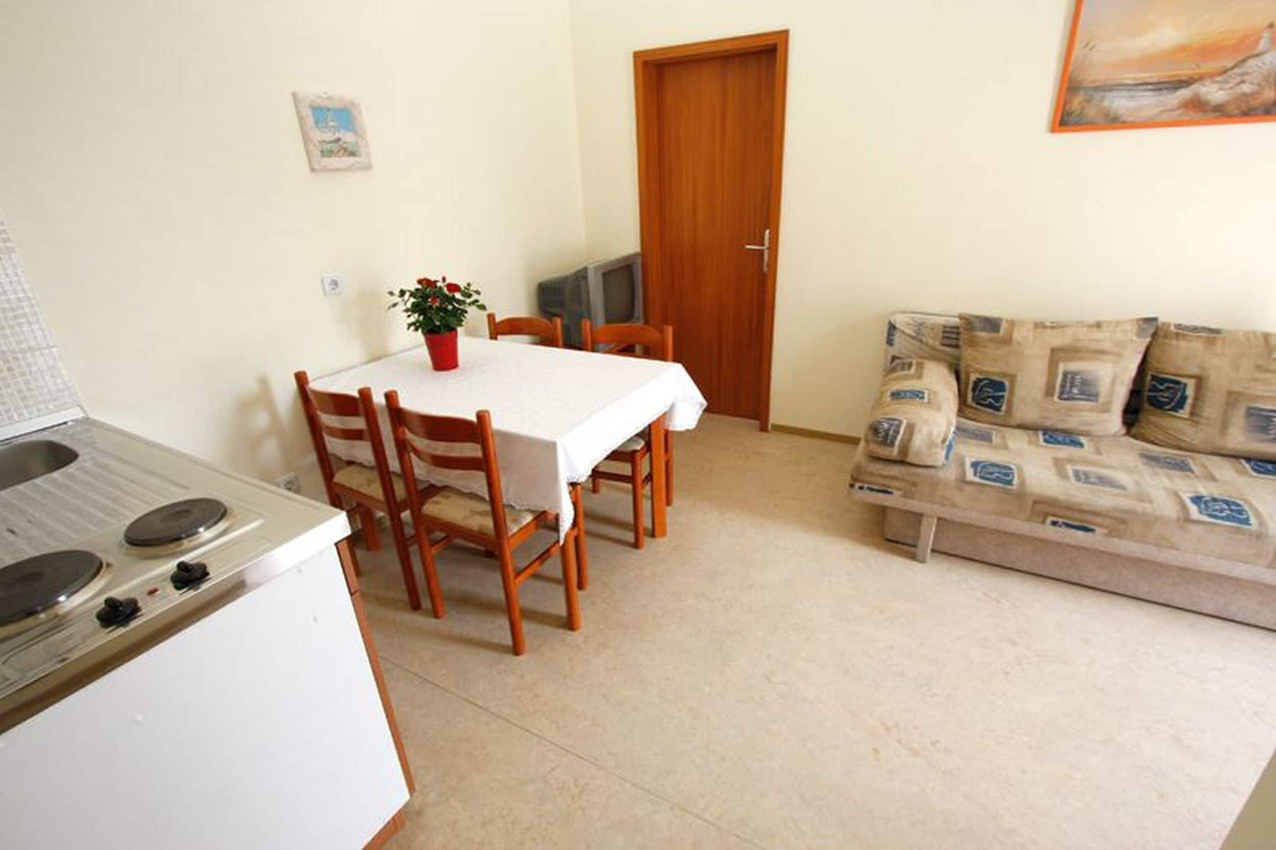 A compact kitchen with a stove, dining table, and chairs. A television is mounted on the wall, and a sofa is placed against the wall.