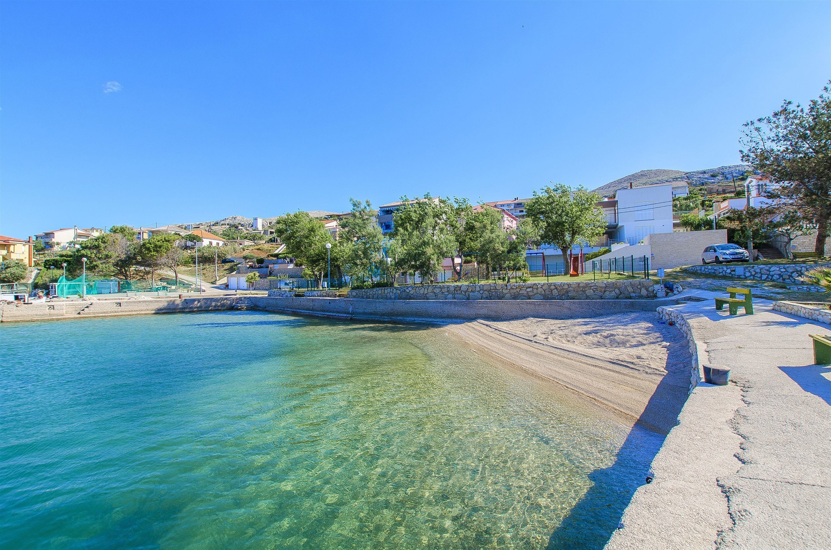 A coastal scene with clear water, a sandy beach, and buildings in the background.