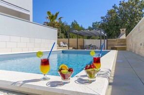 A poolside area with a clear blue pool, two drinks with fruit garnishes, and a lounge chair in the background. - Apartments Villa V (Vir)