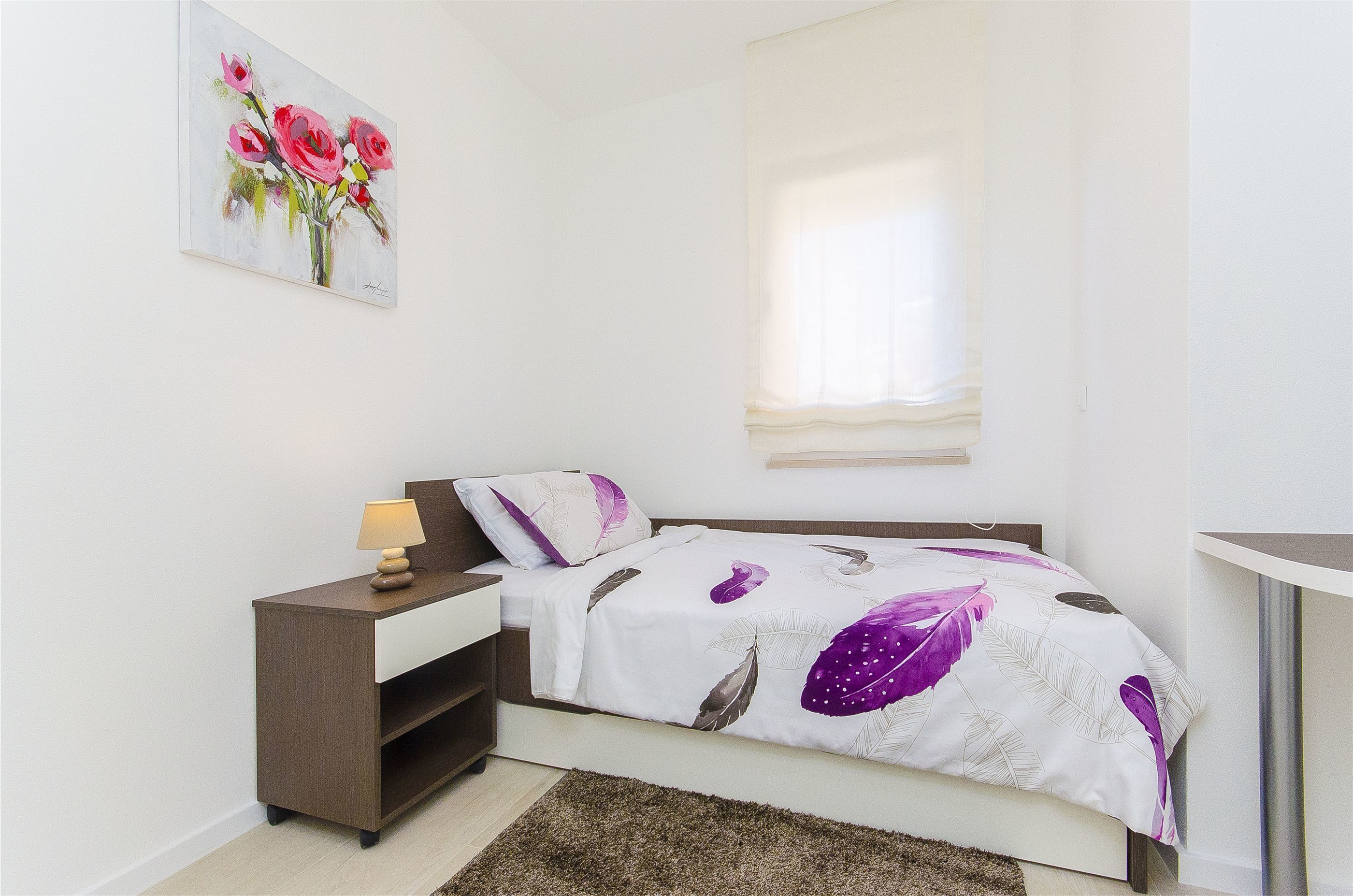 Un dormitorio con cama, mesita de noche con lámpara, mesilla, un cuadro en la pared y una ventana con cortinas.