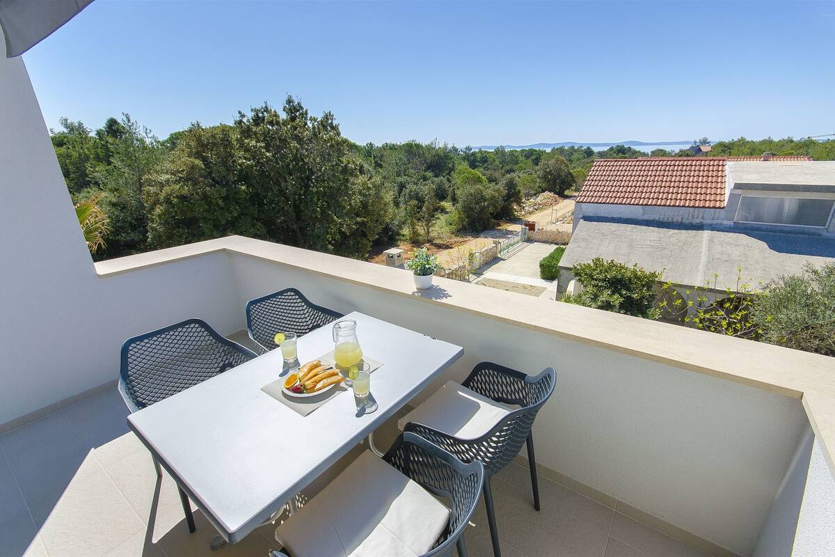 A balcony with a table set for two, chairs, and a view of a green landscape and buildings.