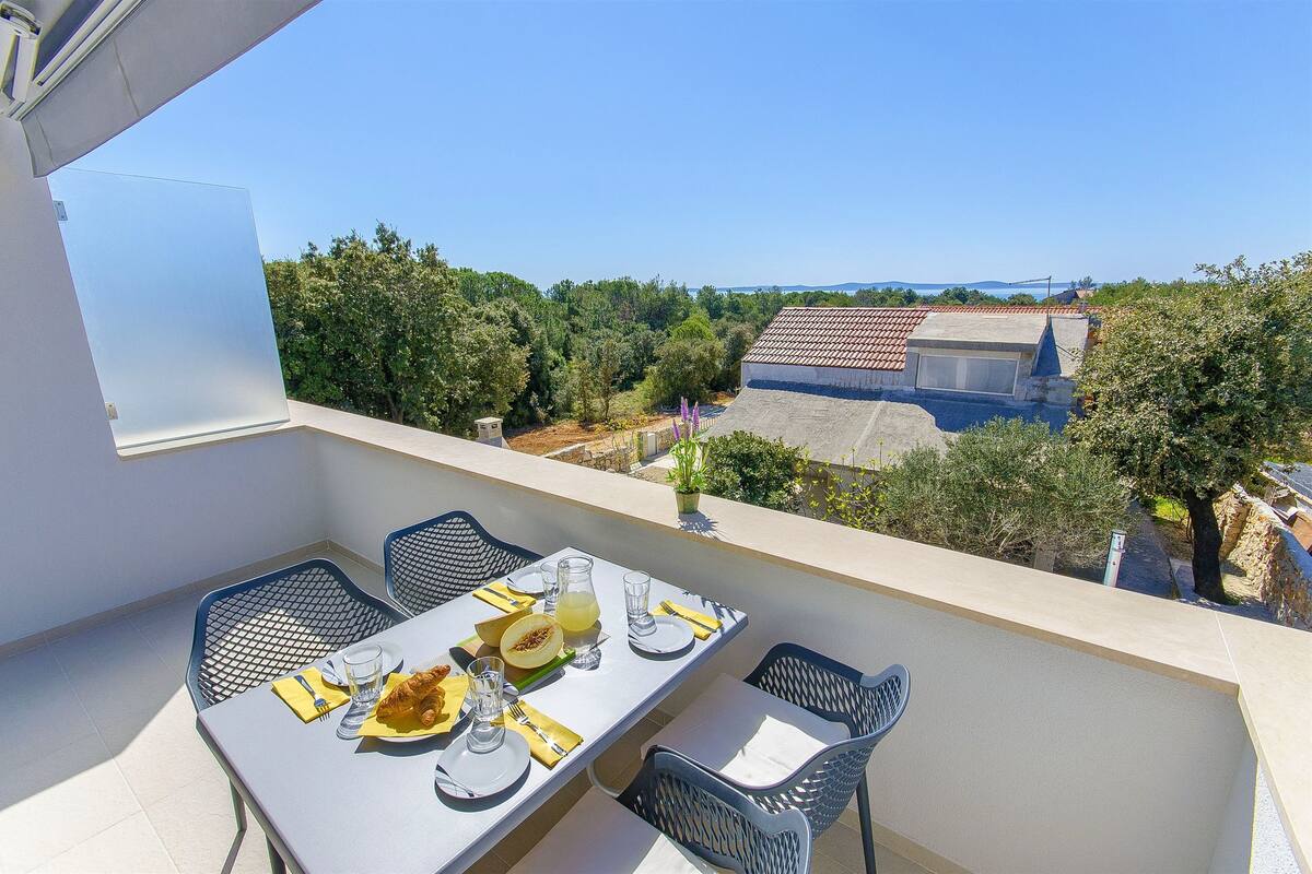 A balcony with a table set for two, overlooking a residential area with trees and a distant view of the sea.