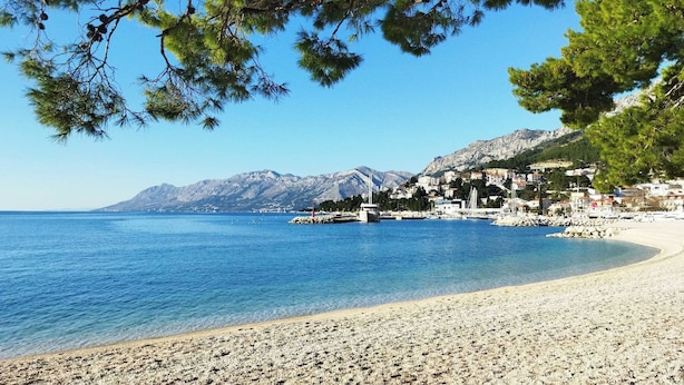 Ein Kiesstrand mit klarem, blauem Meer, eine kleine Stadt mit Häusern an einem Hügel und Berge im Hintergrund.