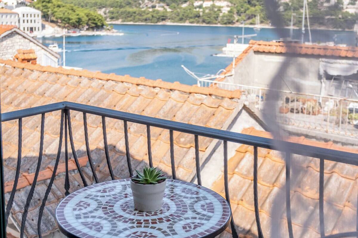 Un balcon avec un sol carrelé, une table ronde agrémentée d’un pot de fleurs, et une vue sur une étendue d’eau et des bâtiments au loin.