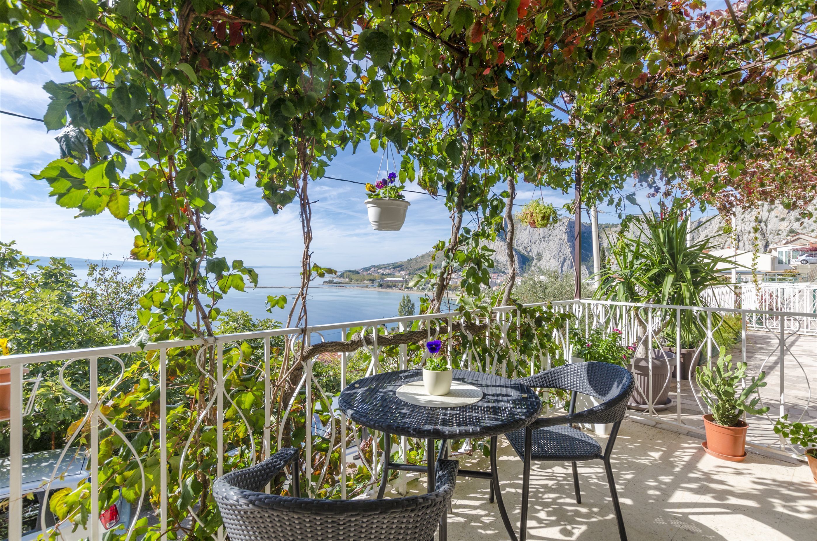En balkon med fletstole og et bord, med udsigt over vand og et bjerglandskab.