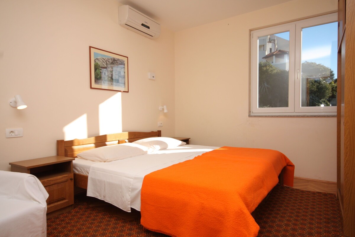 A hotel room with a bed, a nightstand, a bedside table, a window with a view, and a wall-mounted air conditioner.