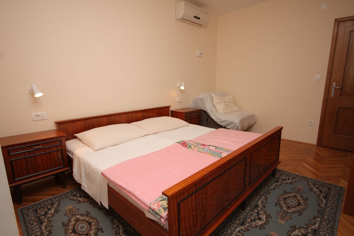 A bedroom with a wooden bed, bedside tables, a nightstand, a chair, an air conditioning unit, and a patterned rug.