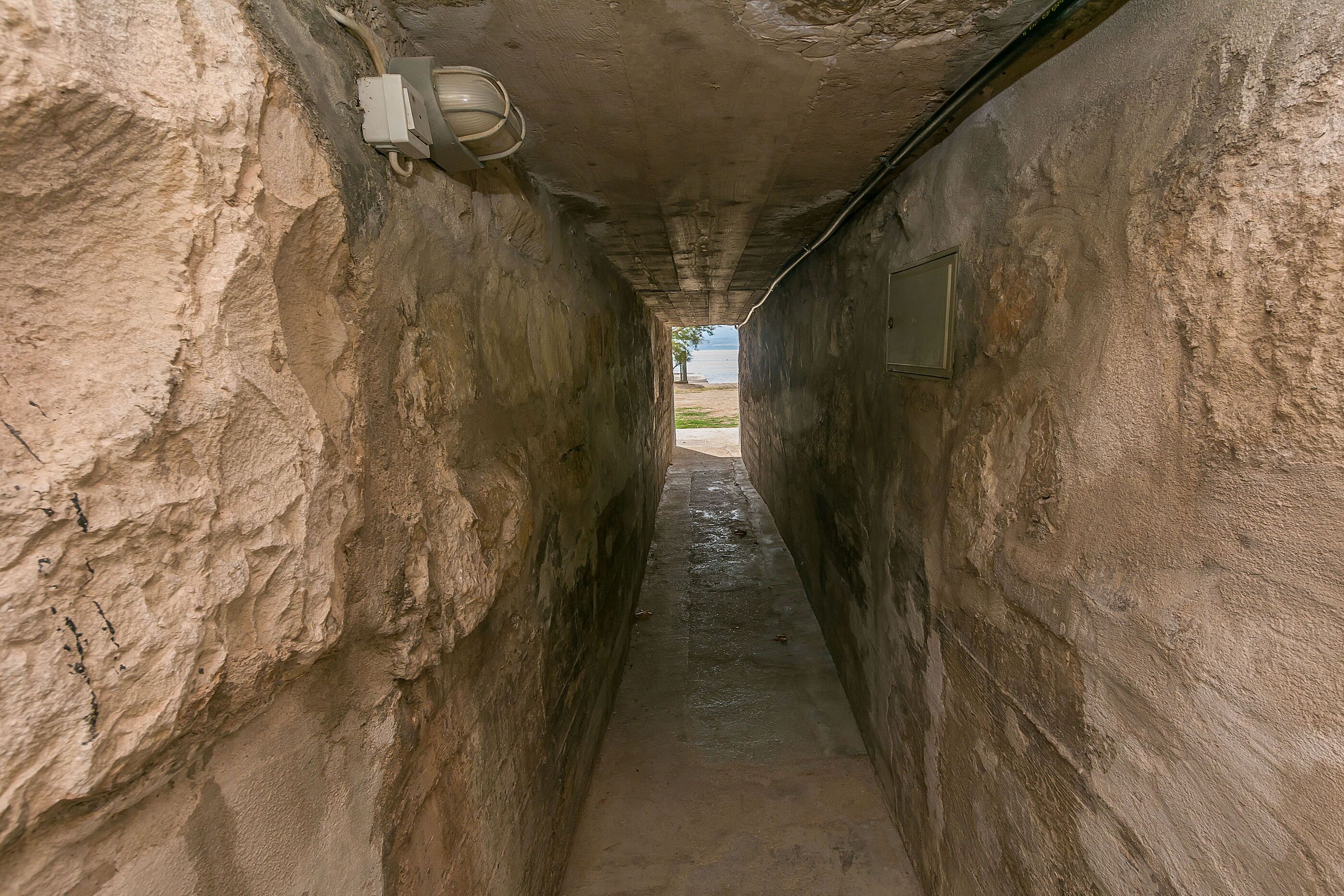 A narrow, stone-walled corridor with a concrete floor, a light fixture, and a door at the end.
