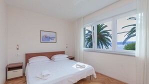 Una cama bien hecha con sábanas blancas, cabecera de madera y una mesita de noche con una lámpara. Un cuadro de una escena costera cuelga en la pared.