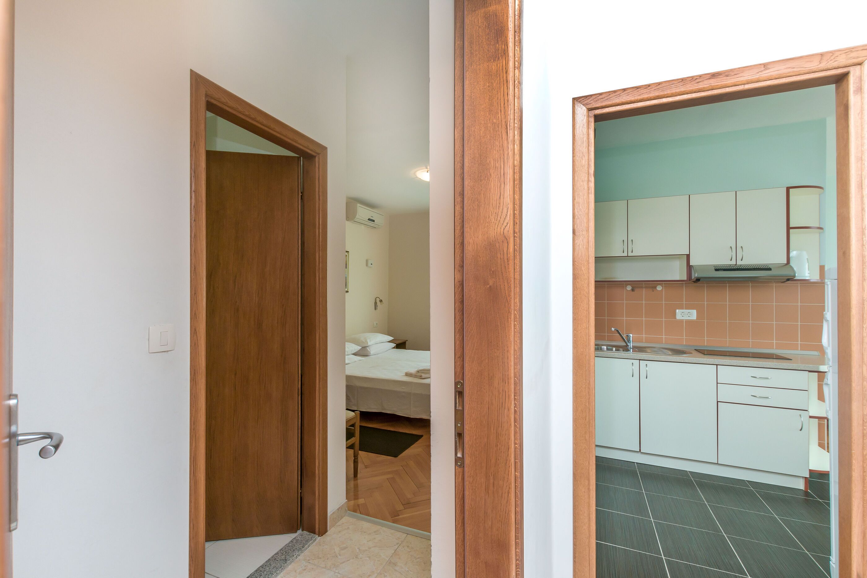 A narrow hallway leading to a bedroom with a wooden door, a small bathroom with a sink, and a kitchenette with white cabinets and a stove.