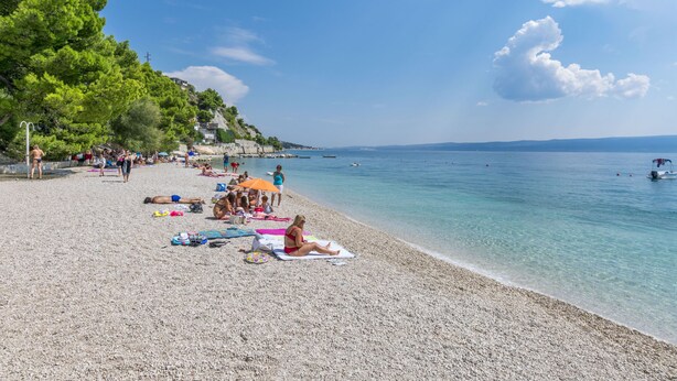 Una playa de guijarros con gente tomando el sol y nadando, agua azul cristalina y una costa lejana con edificios.