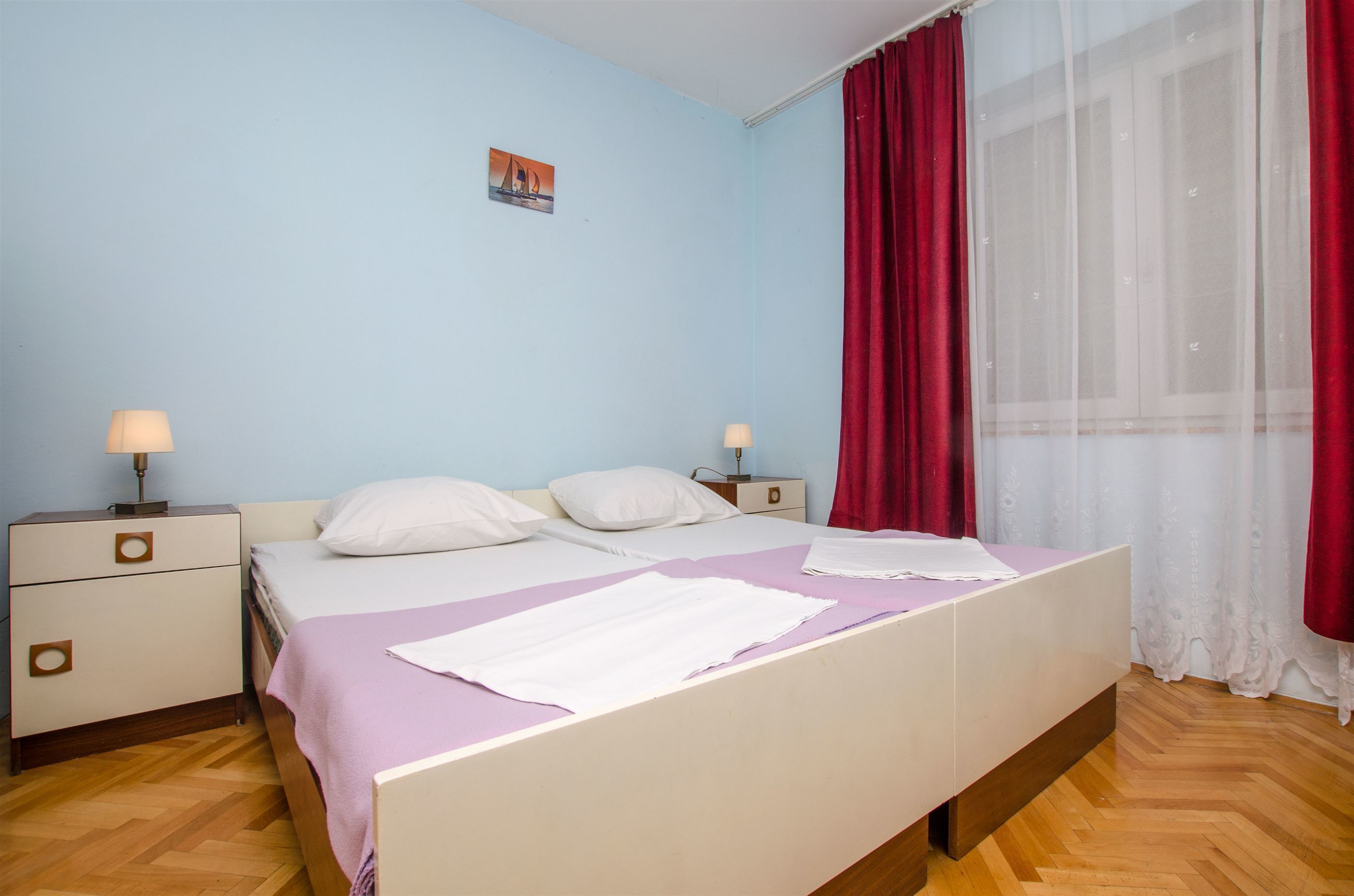 A bedroom with a bed, two bedside tables, a nightstand, a window with curtains, and a wall-mounted picture.