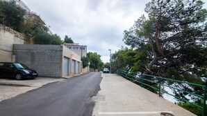 A paved pathway alongside a road with a parked car, a building with a garage, and a railing by a body of water. - Apartments Igor (Bratus)