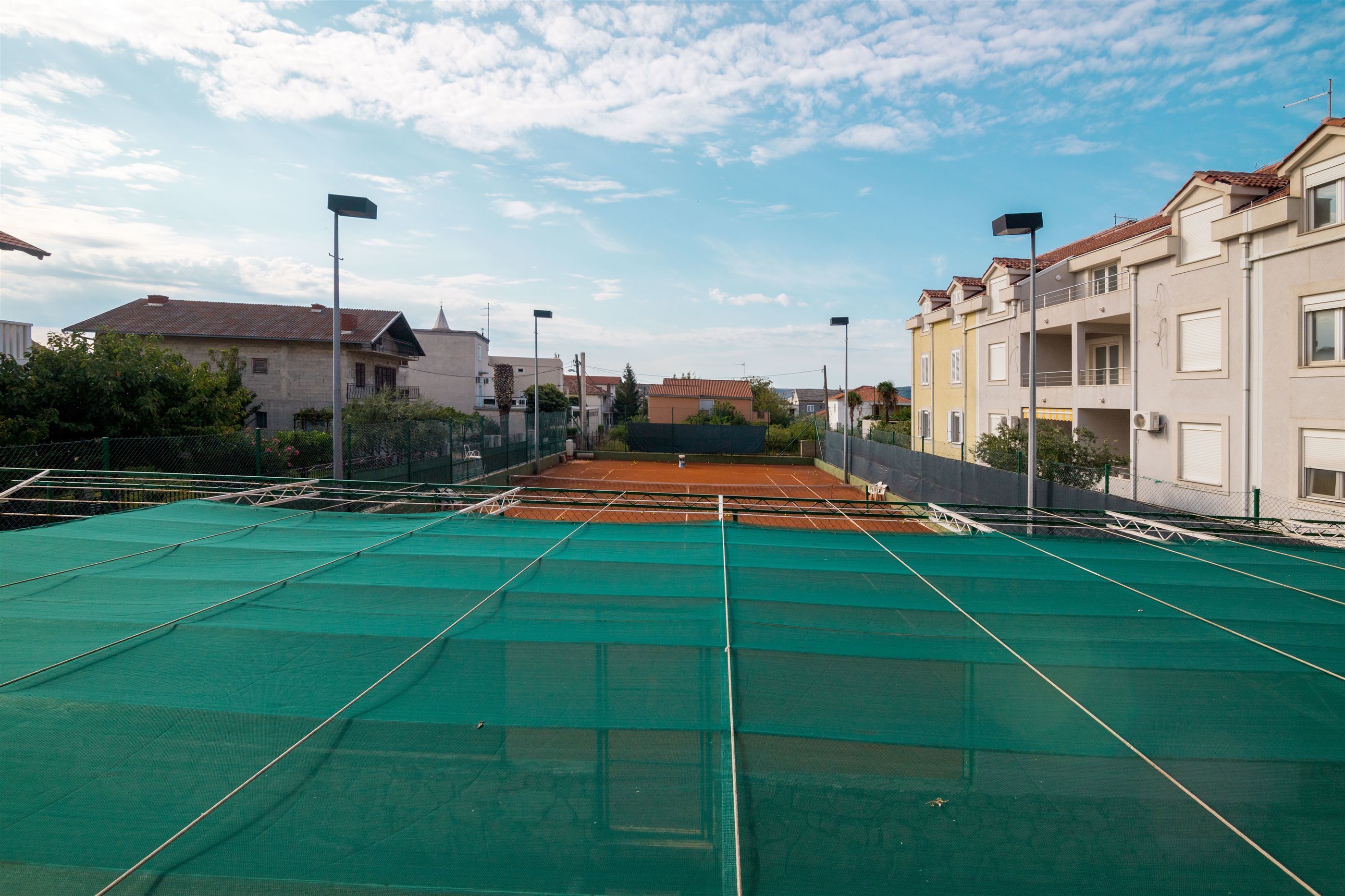 Een tennisbaan met een groene ondergrond, omgeven door woongebouwen en lantaarns.