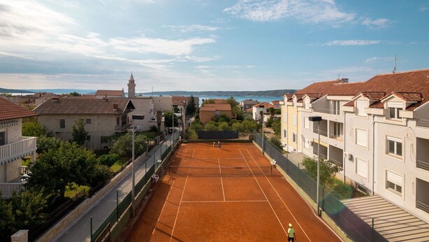 Una cancha de tenis rodeada de edificios residenciales, cielo despejado y una torre al fondo.