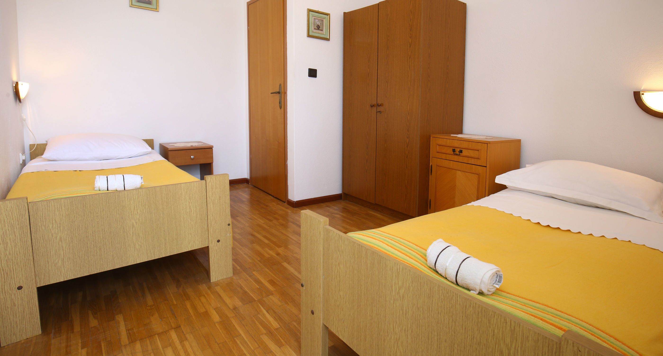A room with two wooden beds, a wardrobe, a nightstand, and a wall-mounted light fixture.