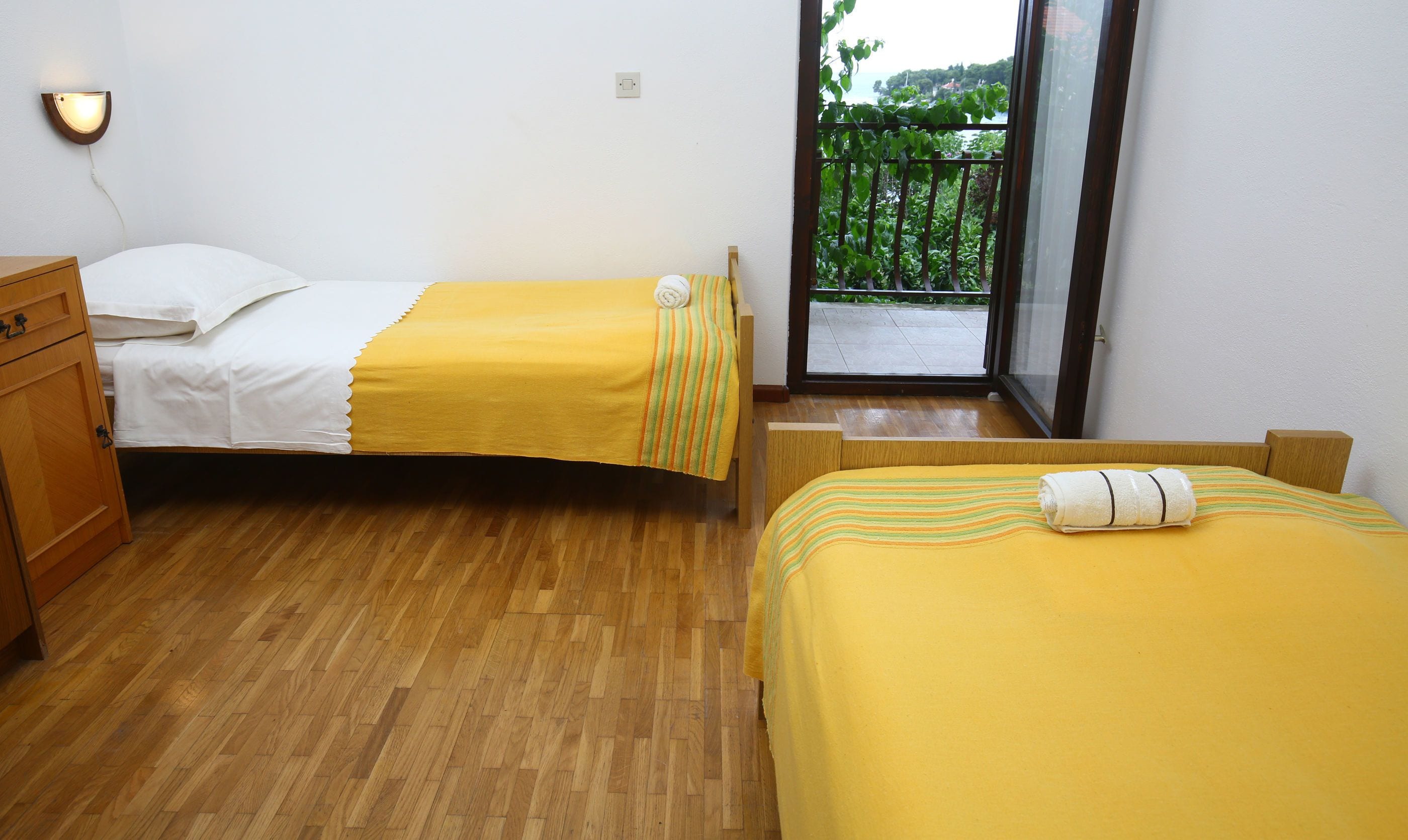 Habitación con dos camas, suelo de madera, un pequeño aparador de madera y un balcón con vistas a la vegetación.