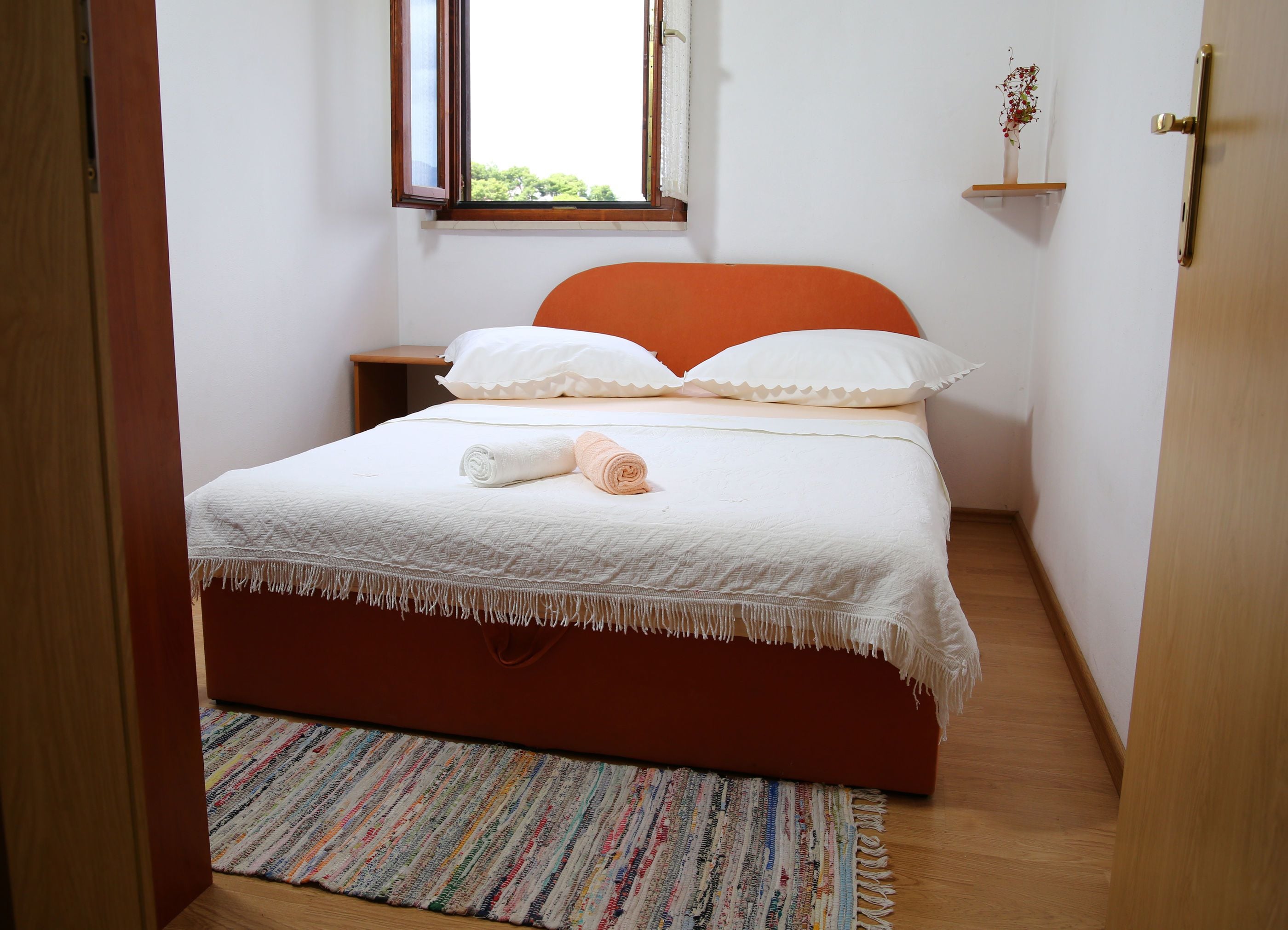A bedroom with a bed, a window, a nightstand, a shelf, and a door.