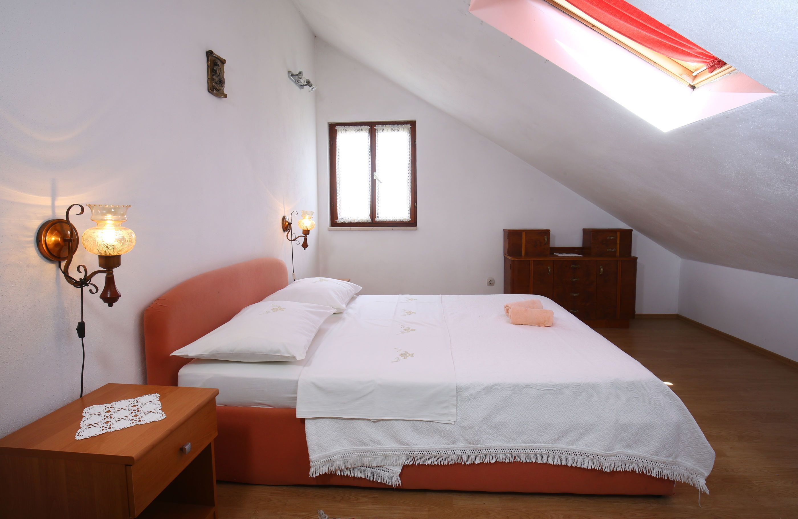 Une chambre avec un plafond en pente, un lit avec une literie blanche, deux lampes fixées au mur, une table de chevet en bois, une petite table et une fenêtre avec un store rouge.