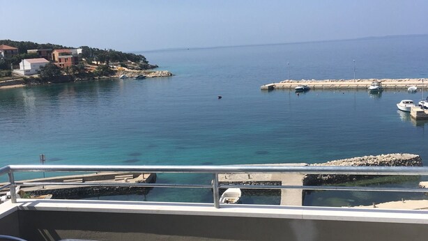 A balcony with a view of a calm sea, a pier with boats, and buildings in the background. - Apartments Suzana (Jakisnica)