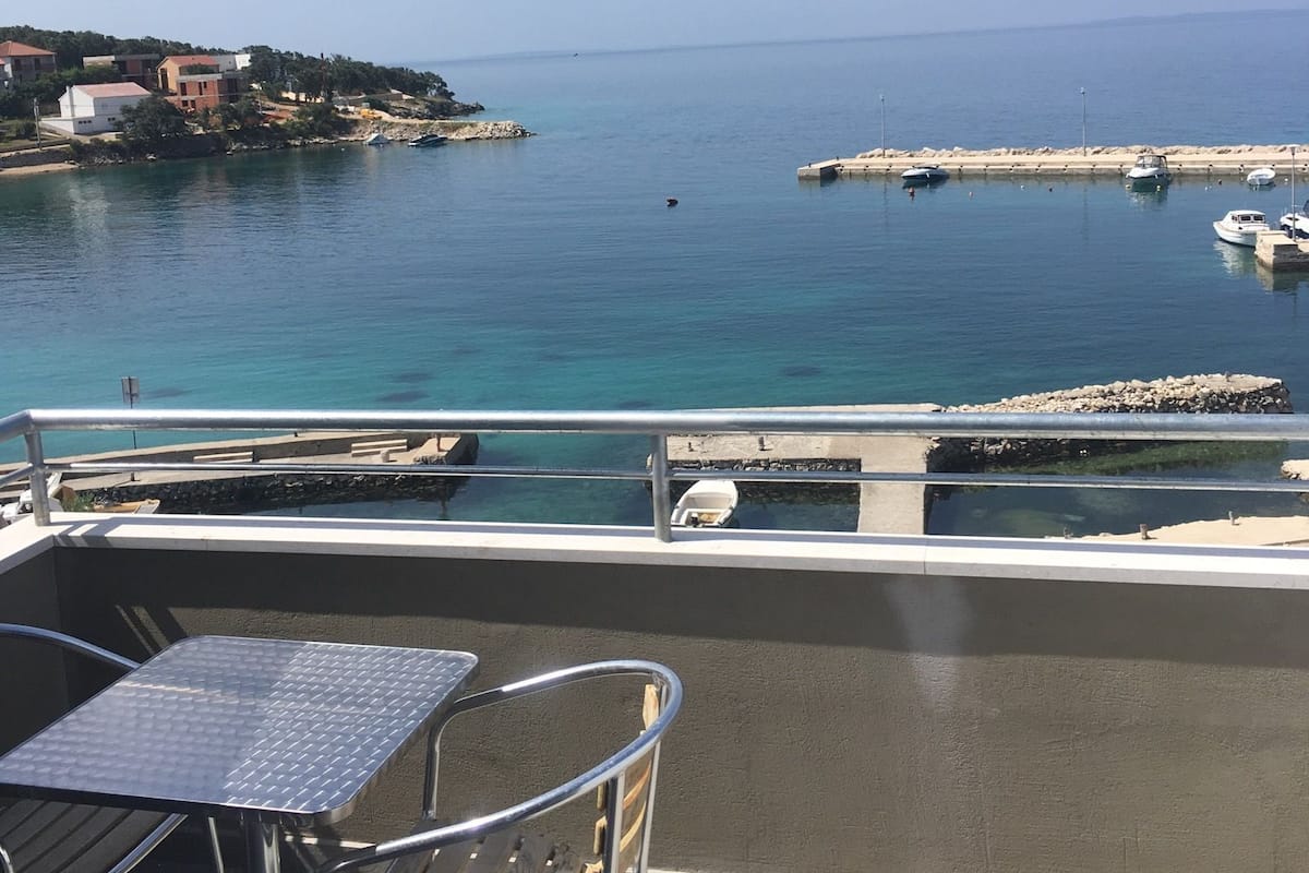A balcony with a view of a calm sea, a pier with boats, and buildings in the background.
