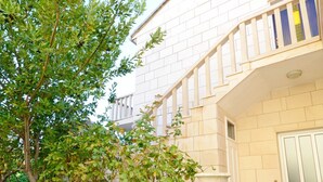 A modern building with a stone facade, a staircase leading to a balcony, and a small garden area with trees and a fountain. - Apartment Jerka (Supetar)