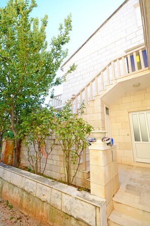 A modern building with a stone facade, a staircase leading to a balcony, and a small garden area with trees and a fountain. - Apartment Jerka (Supetar)