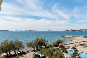 Egy tengerparti kilátás, ahol tiszta az ég, fák, parkoló autók és egy strand látható.