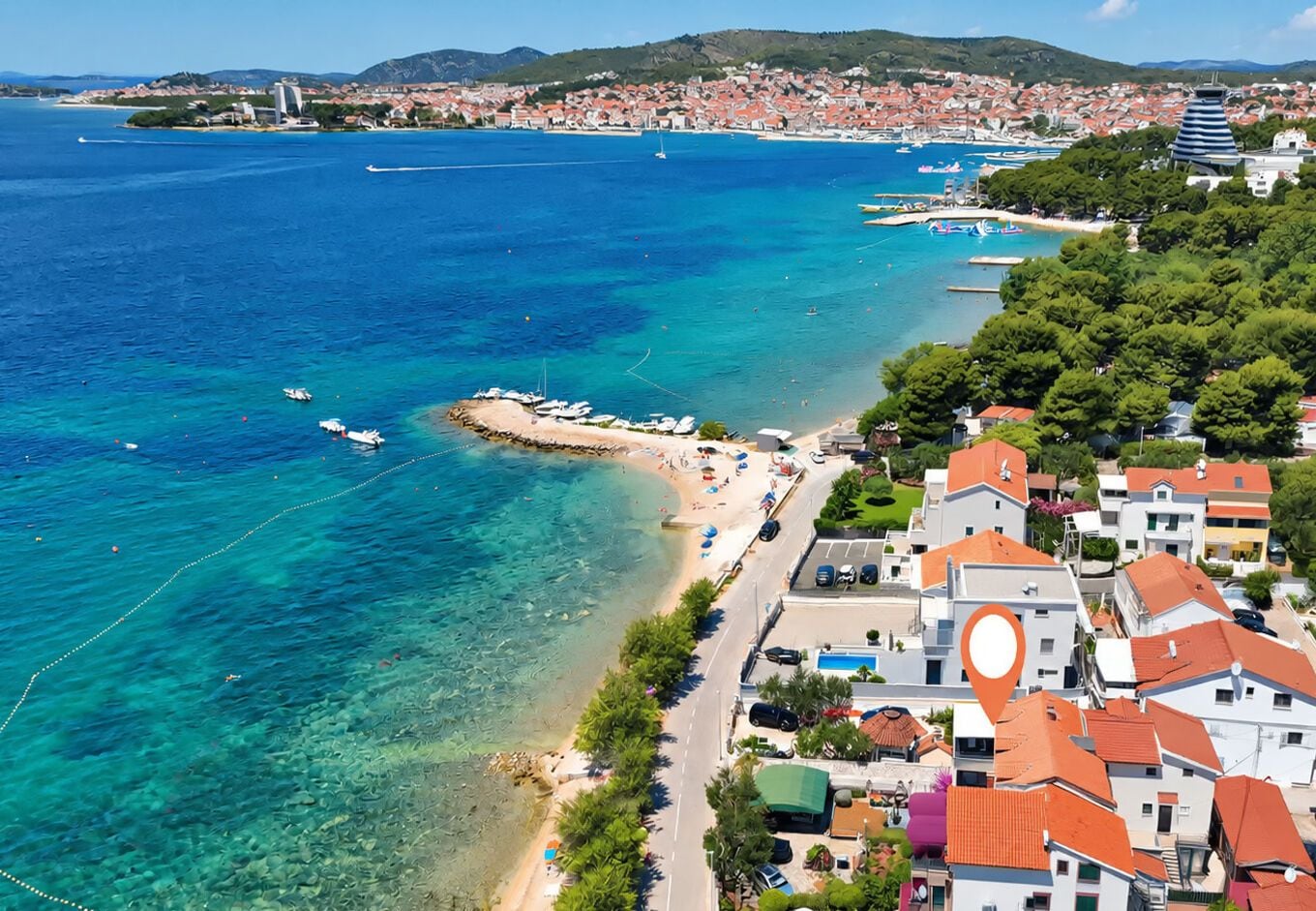 A coastal town with clear blue waters, a sandy beach, and residential buildings with red roofs.