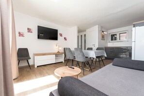 Egy modern, jól megvilágított szoba, melyben síkképernyős televízió, szürke színű, kárpitozott bútorok, egy fa kávézóasztal és egy étkező található, ahol asztal és székek állnak.