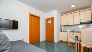 Ein kompakter Wohnraum mit Sofa, Esstisch mit Stühlen, Küchenzeile und einem an der Wand befestigten Fernseher.