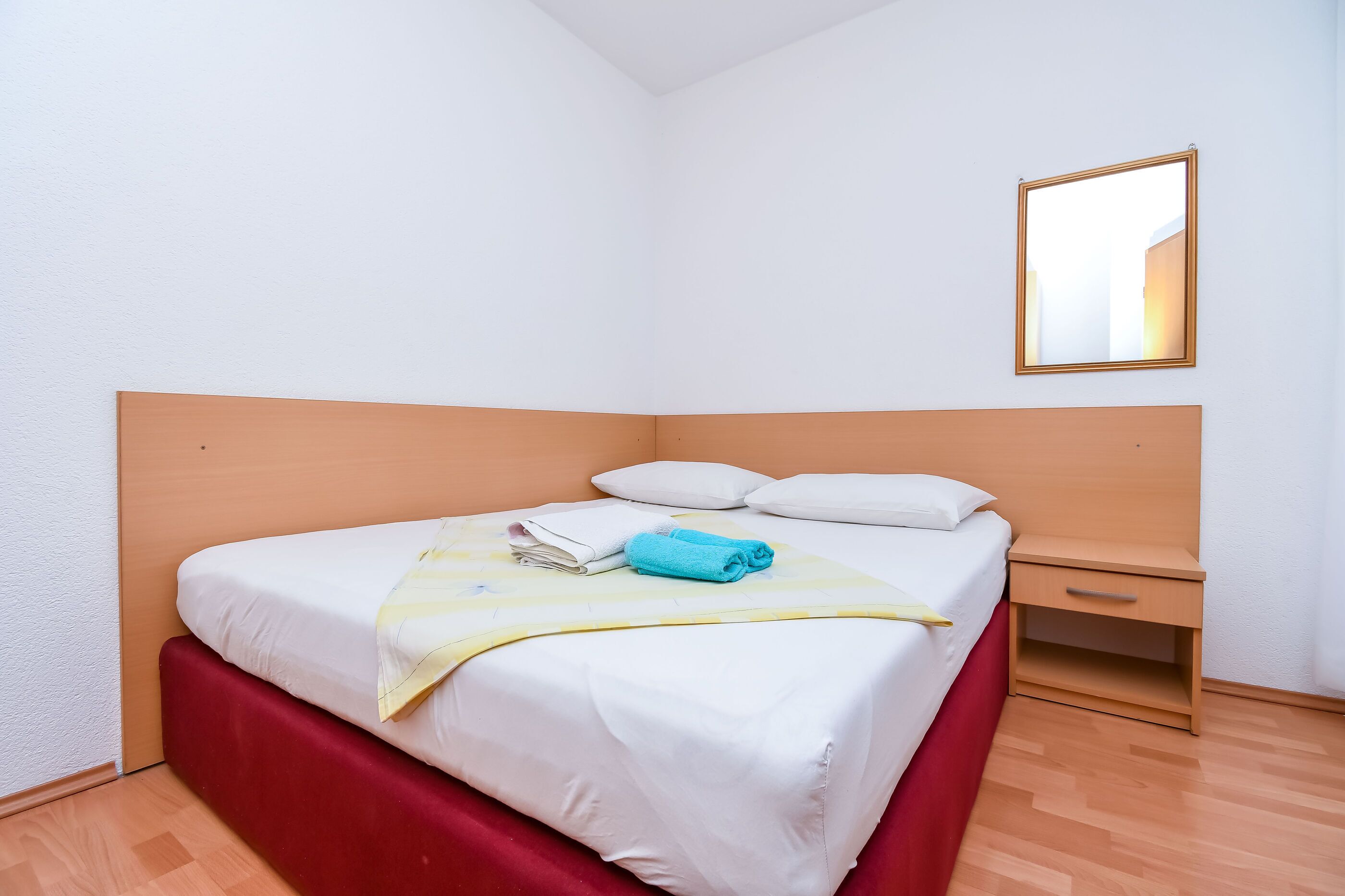 Ein ordentlich bezogenes Bett mit weißer Bettwäsche und einem roten Kopfteil, ein Nachttisch mit Schublade sowie ein Spiegel an der Wand.