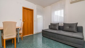 Ein Zimmer mit Holztür, grauem Sofa mit Kissen, Esstisch mit Stühlen und grün gefliestem Boden.