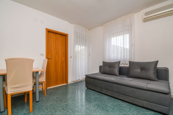 A room with a wooden door, a grey sofa with cushions, a dining table with chairs, and a green tiled floor. - Apartments Jakov (Marina)