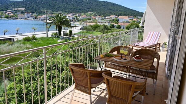 A balcony with wicker furniture, a table with drinks, and a view of a coastal town and mountains. - Apartment Natasa (Vinisce)