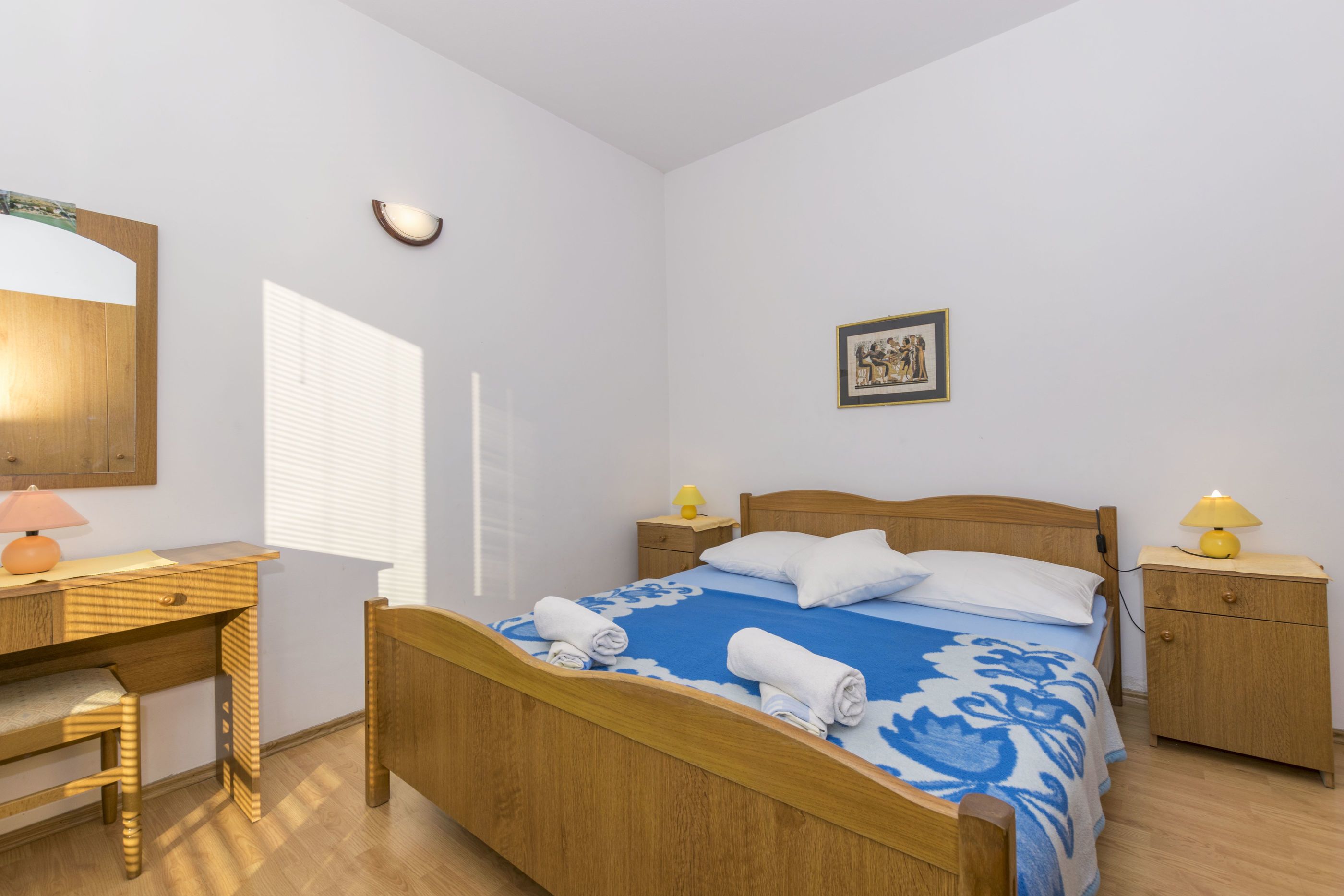 A bedroom with a wooden bed, a desk, a chair, a nightstand with a lamp, and a framed picture on the wall.
