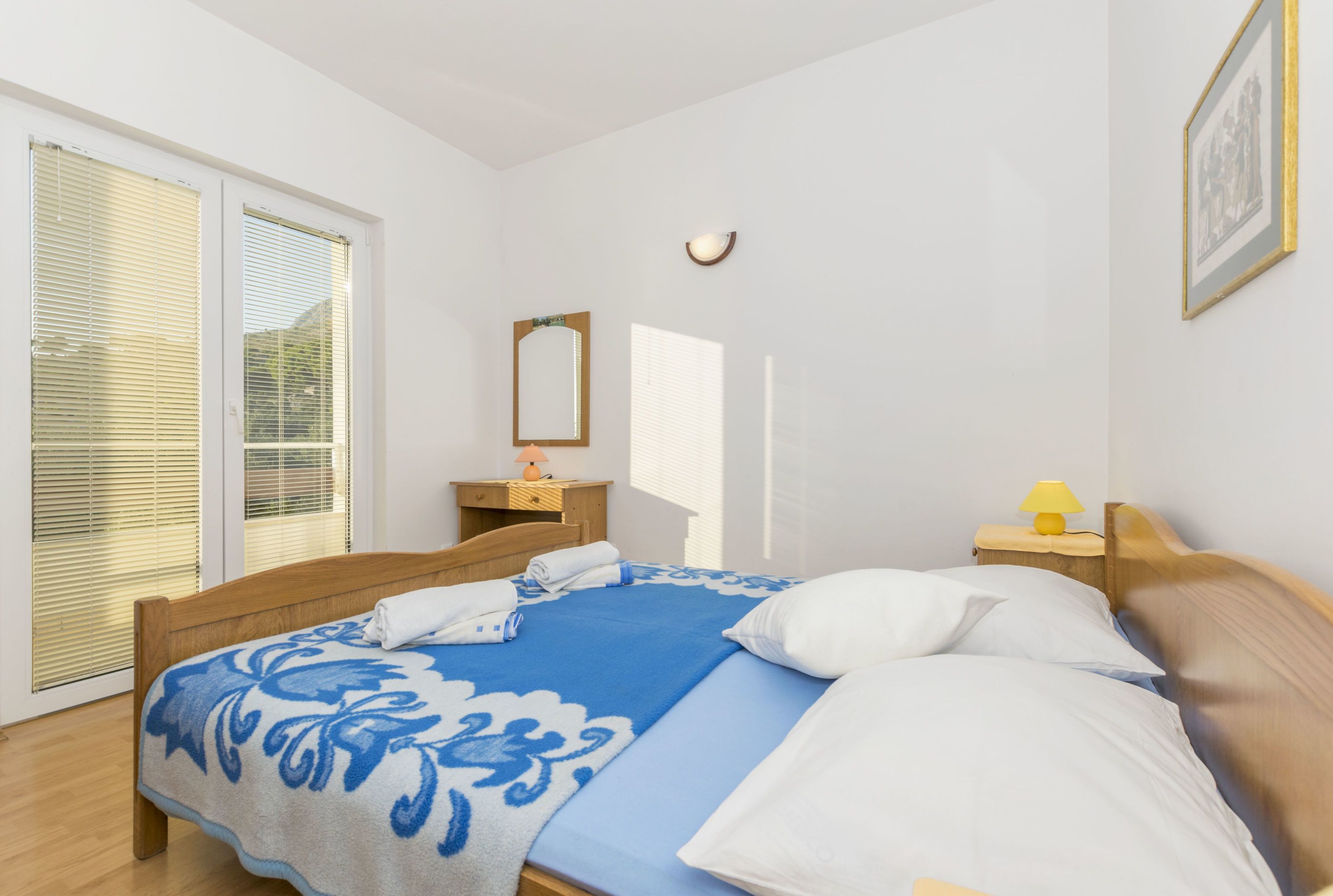 Een slaapkamer met een bed, een nachtkastje, een lamp, een spiegel en een raam met zonwering.