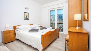 A bedroom with a wooden bed, a nightstand, a desk, and a chair. There is a window with a view of mountains and a door leading to a balcony.