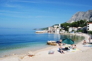 A beach with clear water, a rocky outcrop, and buildings in the background.