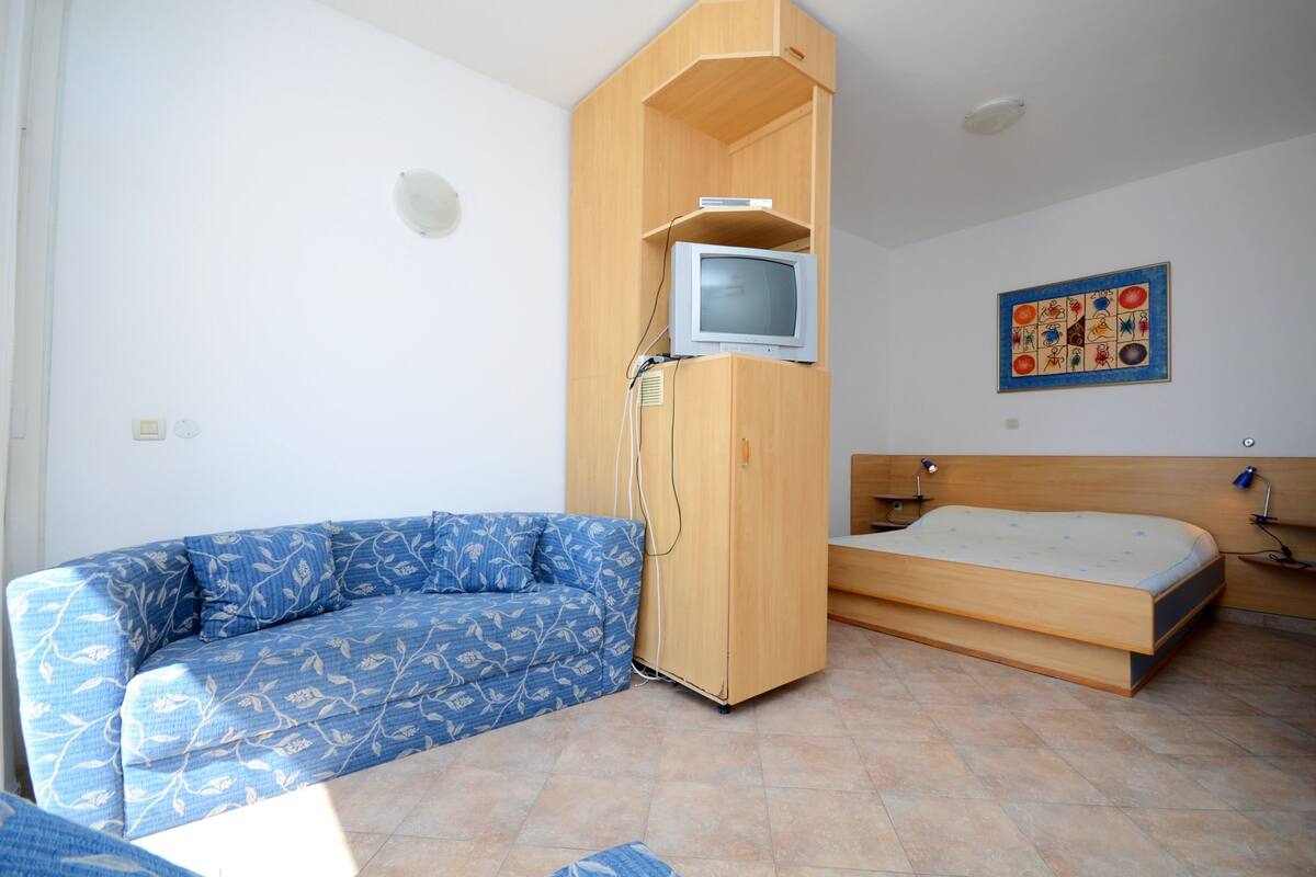 A small room with a blue patterned sofa, a wooden wardrobe with a television on top, a bed with a wooden headboard, and a wall decoration.