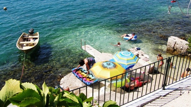 A clear blue sea with a boat, people in inflatable rings, and a colorful umbrella on a rocky shore. - Apartment Zeljka (Seget Vranjica)