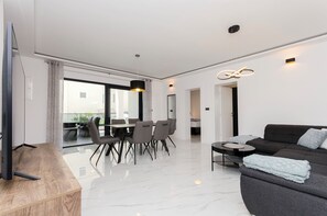 Ein modernes Wohnzimmer mit Fernseher, Essbereich mit Tisch und Stühlen, Sofa mit Decke und ein großes Fenster mit Vorhängen.