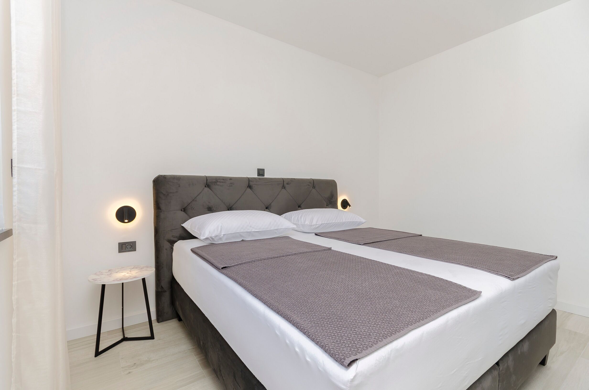 A neatly made bed with a grey headboard and white bedding, a small bedside table, and a wall-mounted light fixture.