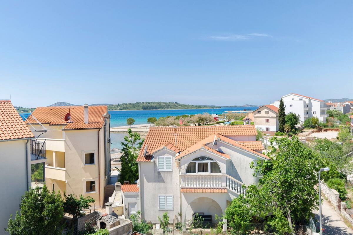 Zona residencial costera con edificios de tejados de teja, mar azul cristalino y un paisaje montañoso al fondo.