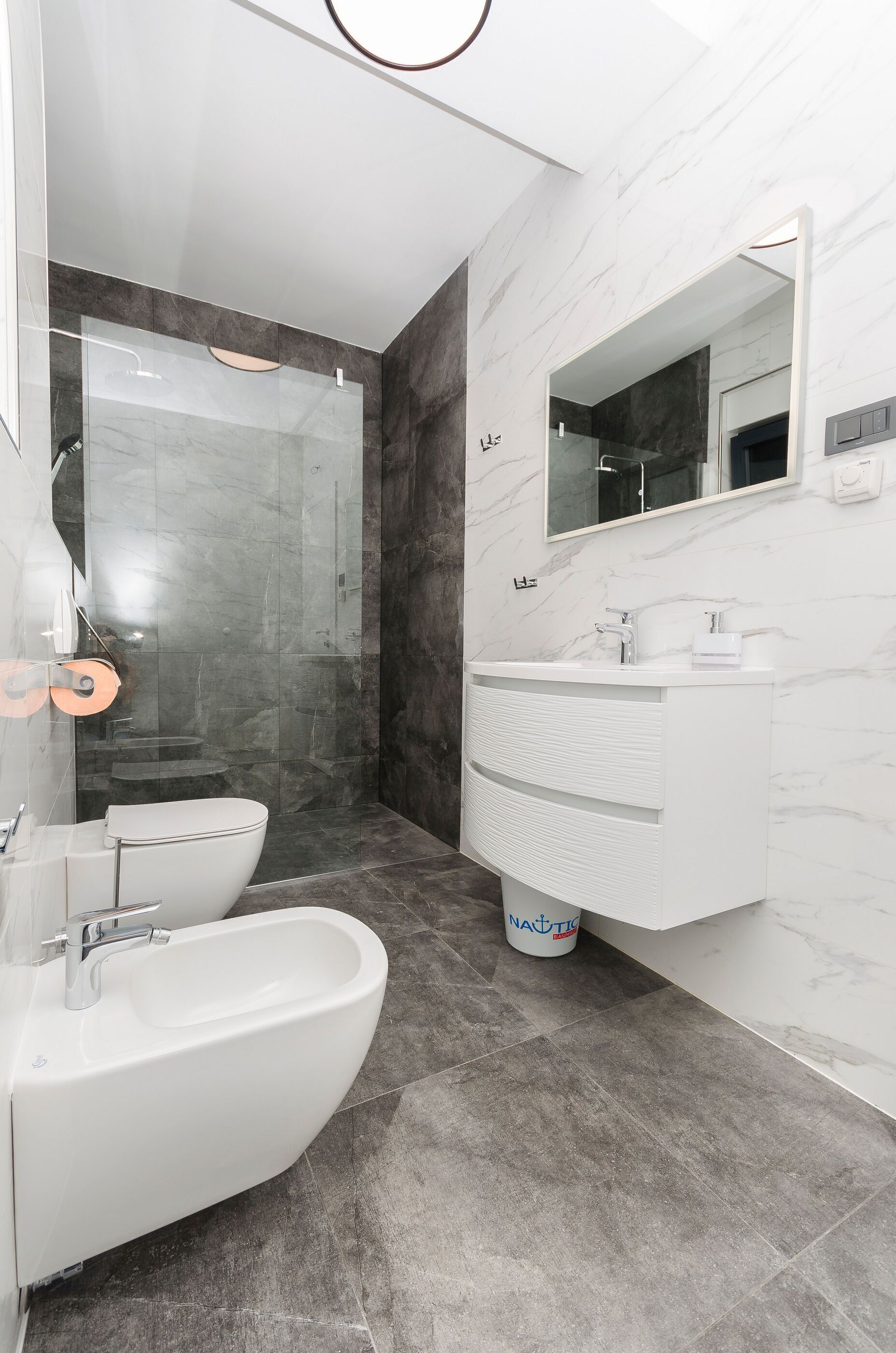 Un bagno moderno con cabina doccia in vetro, lavandino bianco con piano di lavoro in marmo e un grande specchio sopra il lavandino.