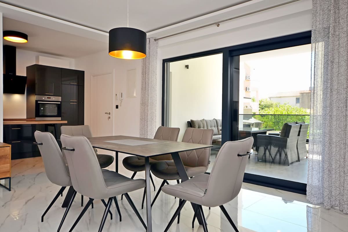 Un comedor moderno con una mesa grande y sillas, una cocina al fondo, y vistas a una zona de estar exterior a través de un ventanal.